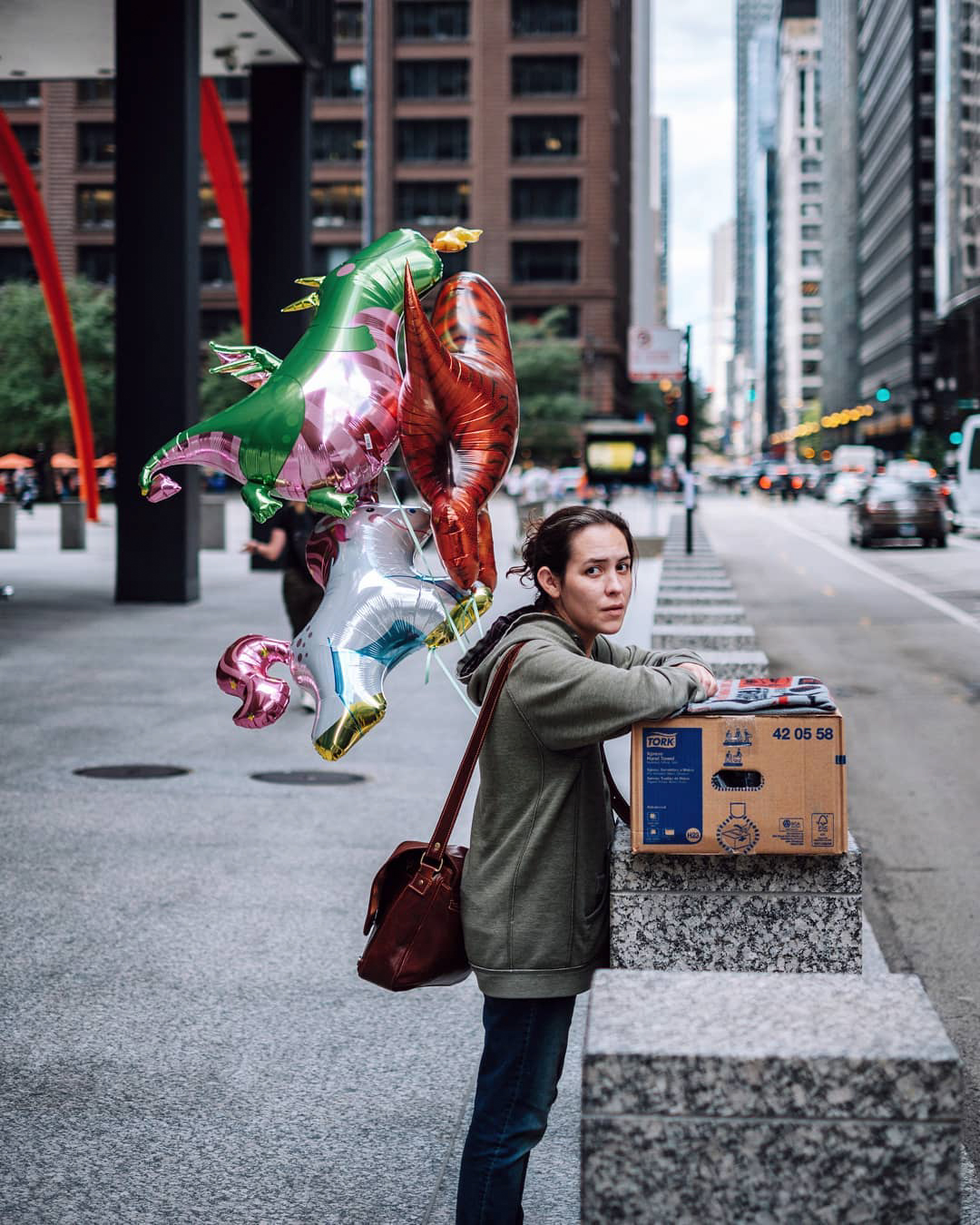 Woman standing near a cardboard box with some balloons on a city street.