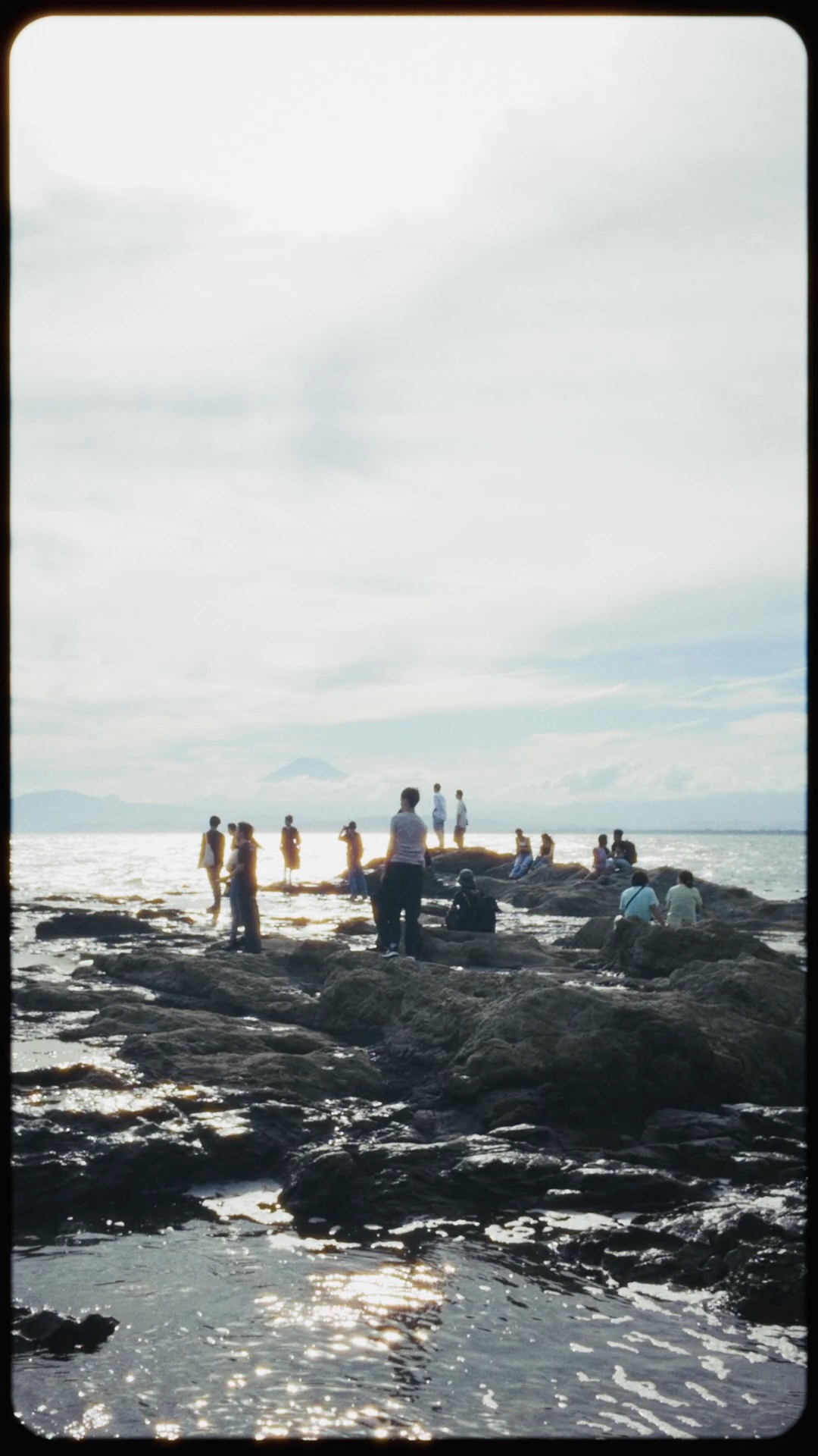 A group of people sit on rocks surrounded by water, with the silhouette of Mount Fuji in the background.